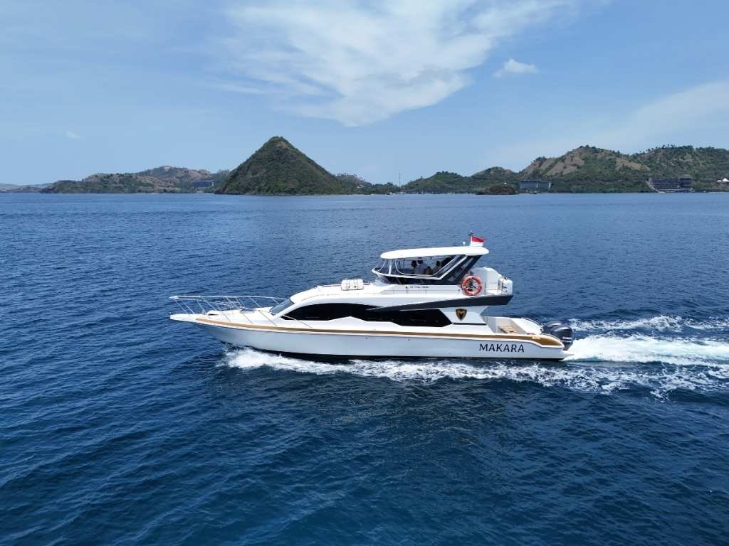 Speed Yacht Makara berwarna putih berlayar di perairan biru Labuan Bajo dengan latar belakang perbukitan hijau. Kapal ini memiliki desain flybridge dan kabin tertutup mewah, ideal untuk Private Charter Labuan Bajo yang eksklusif.